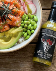 Bowl of sushi with salmon, avocado, and radish next to a bottle of 'Sauce Boss Cake' hot sauce on a wooden surface.