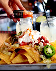 Stack of nachos with various toppings on a tray, with a hand holding a bottle of hot sauce in the background.