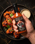Hand holding a bottle of hot sauce over a plate of grilled chicken with a side of dipping sauce on a dark marble surface.