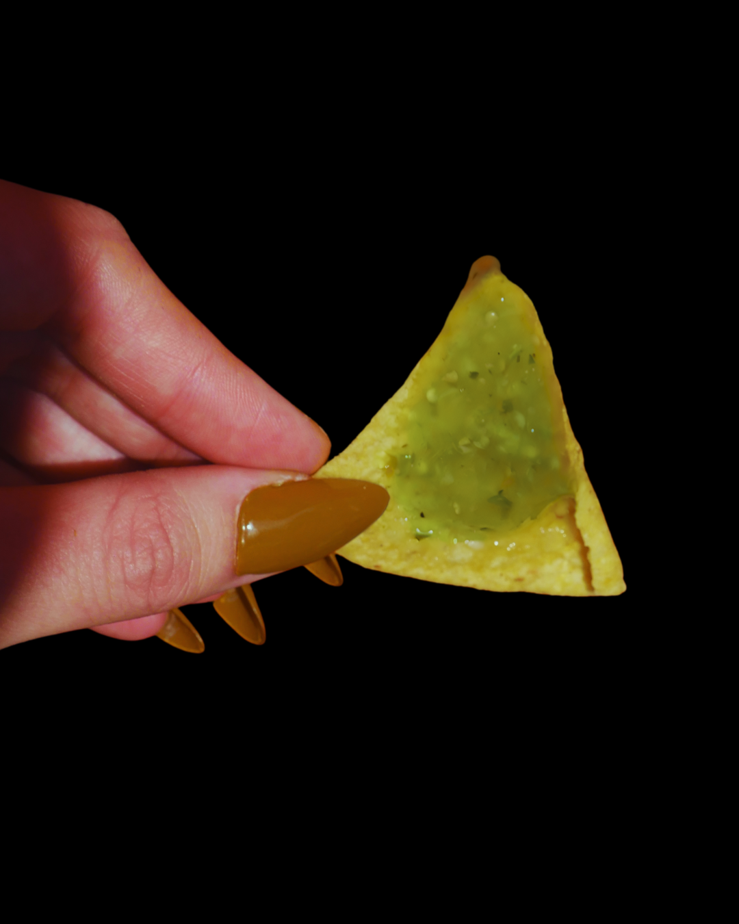 Hand holding a triangular tortilla chip with green sauce against a black background