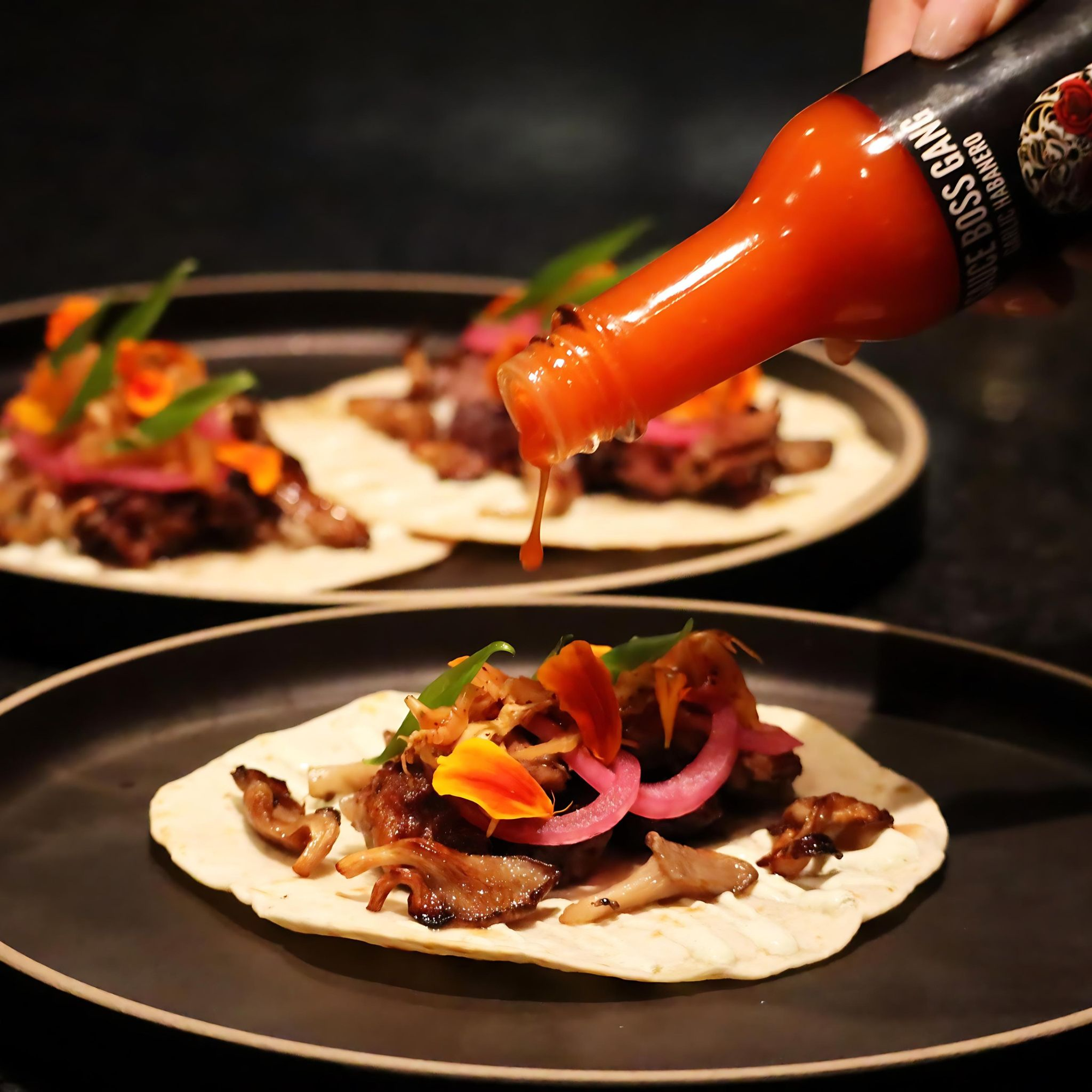 Person pouring hot sauce onto a taco with various toppings on a dark surface.