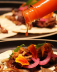 Person pouring hot sauce onto a taco with various toppings on a dark surface.