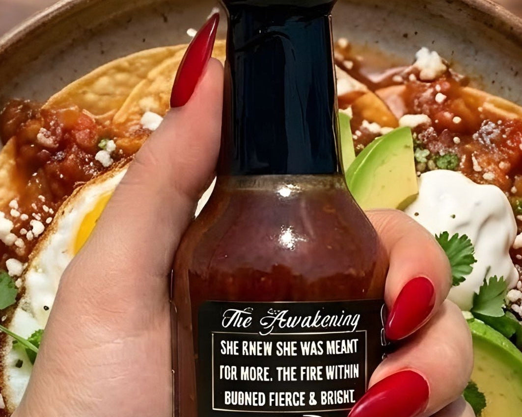 Hand holding a bottle of hot sauce over a plate of food on a wooden table.