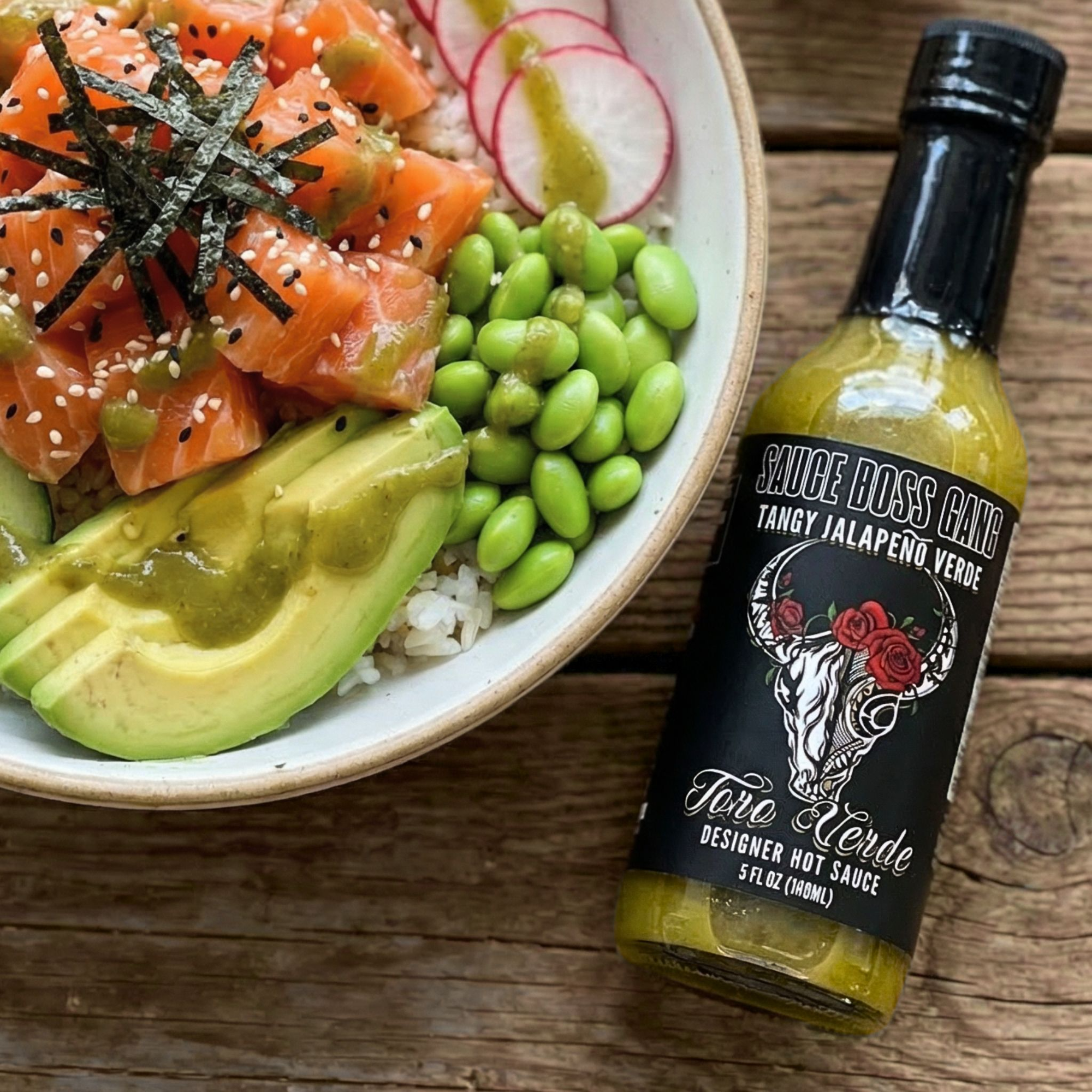 Bowl of sushi with salmon, avocado, and radish next to a bottle of 'Sauce Boss Cake' hot sauce on a wooden surface.