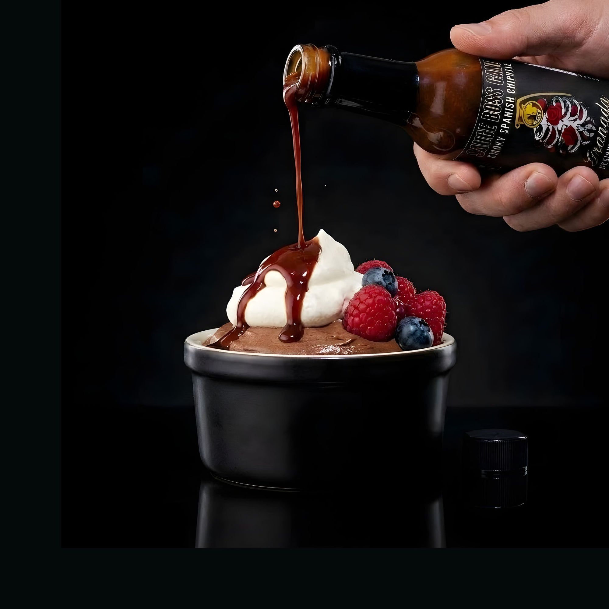Chipotle hot  sauce being poured over a dessert with berries on a black background.