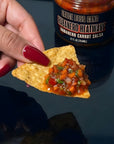 Person holding a chip with salsa next to a jar of Habanero Heatwave salsa.