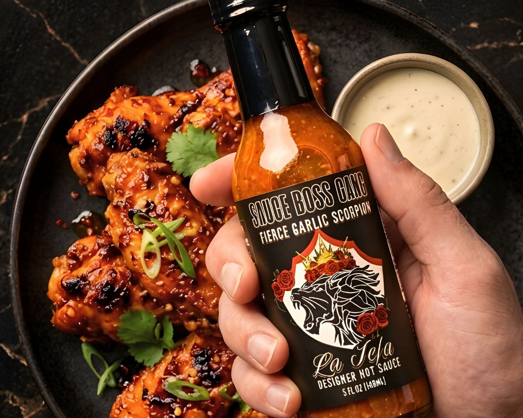 Hand holding a bottle of 'Sauce Boss Gang' hot sauce over a plate of grilled chicken with a side of dipping sauce.