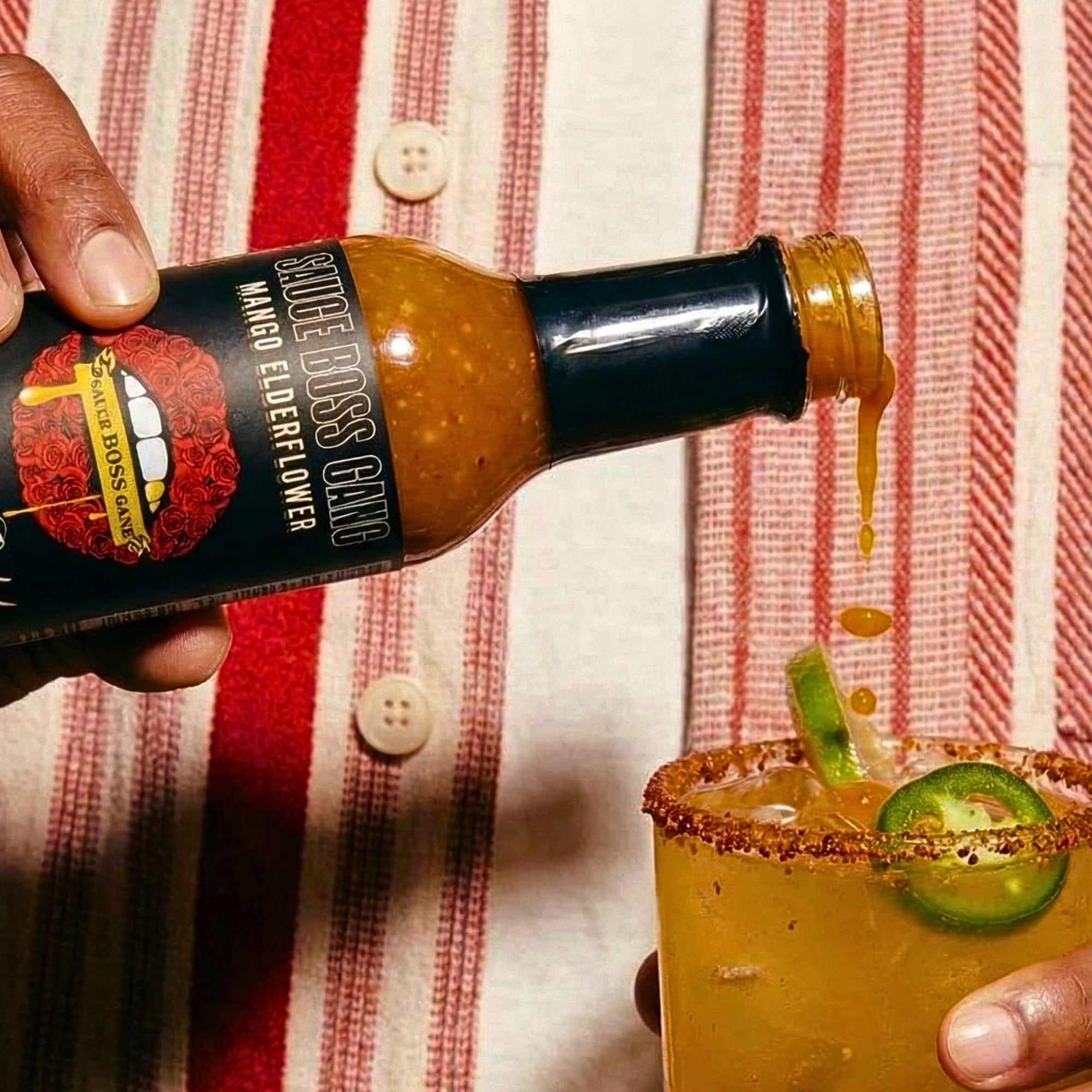 Hand pouring hot sauce into a margarita with a red and white striped shirt in the background