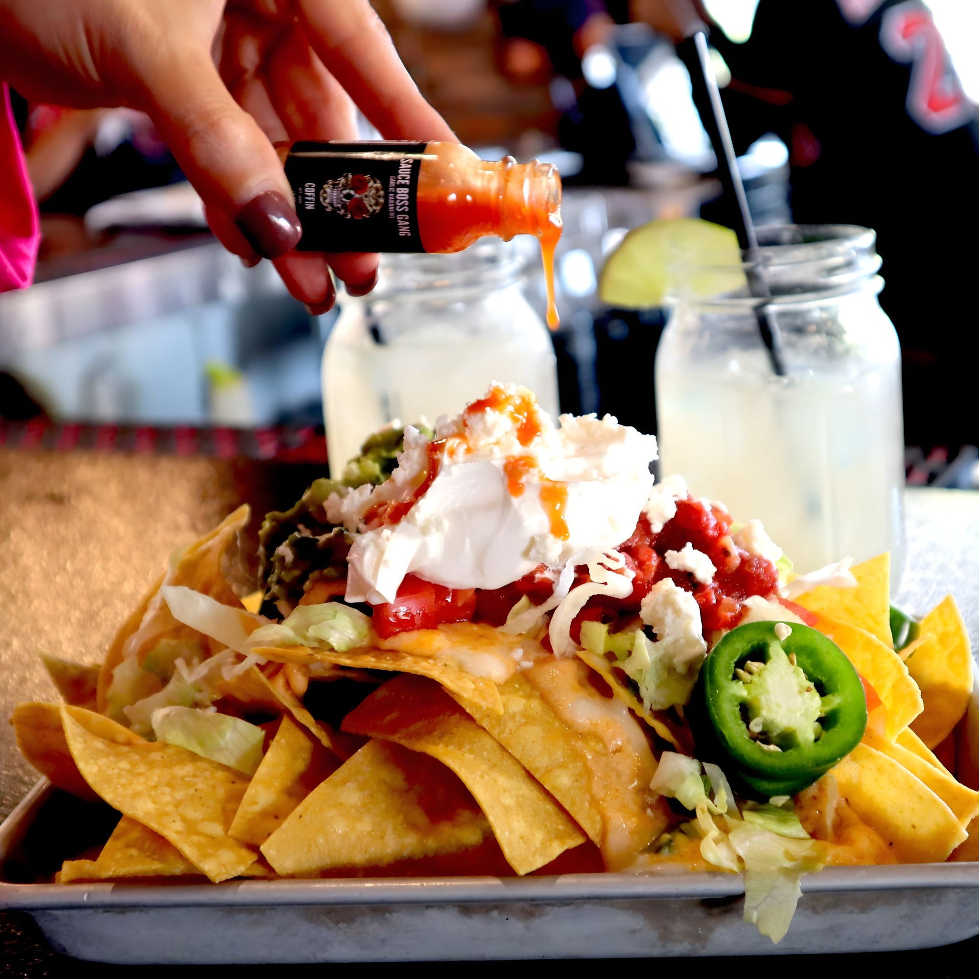 Stack of nachos with various toppings on a tray, with a hand holding a bottle of hot sauce in the background.