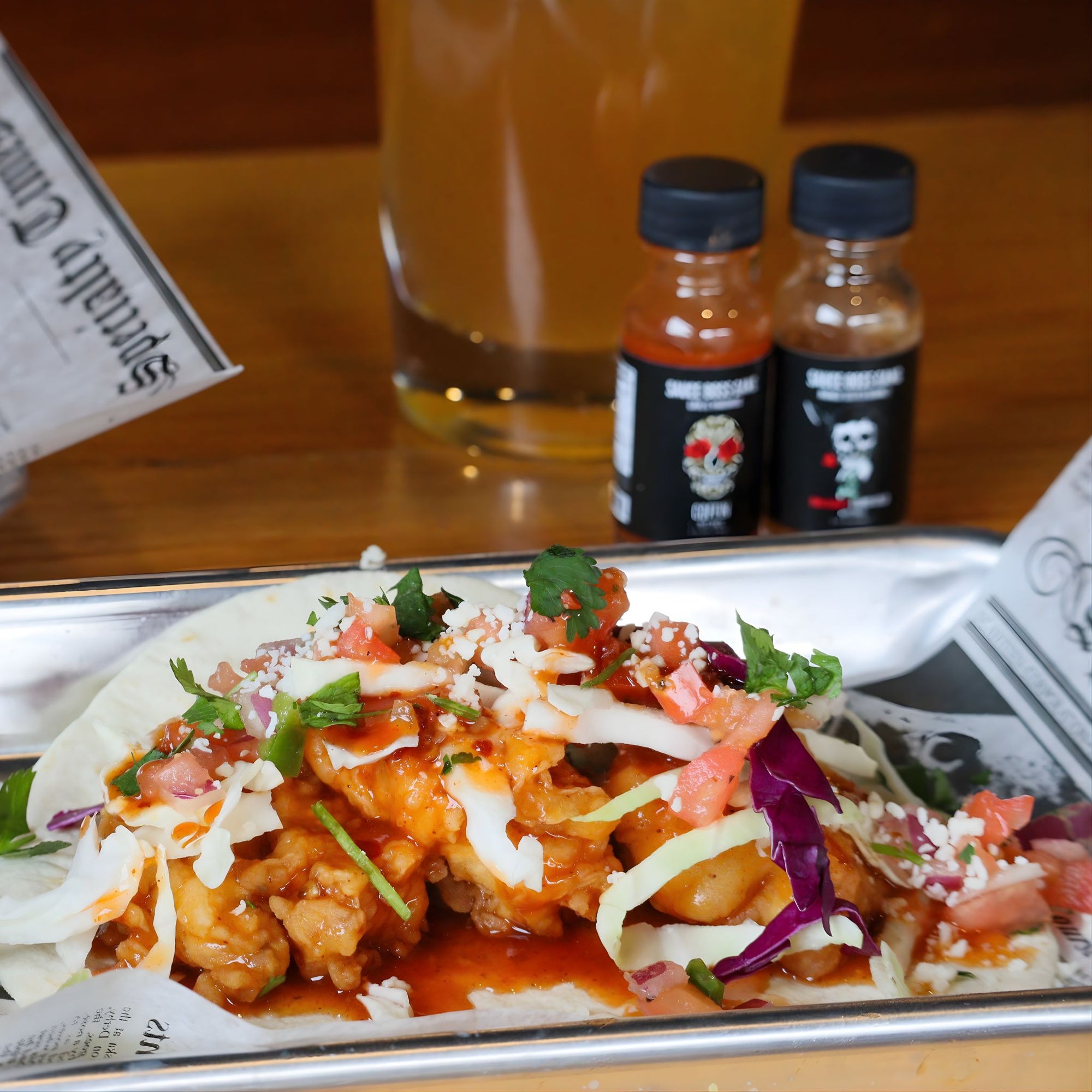 Taco with various toppings on a tray with a glass of beer and two bottles in the background.