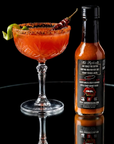 Bottle of hot sauce next to a cocktail with garnishes on a reflective surface
