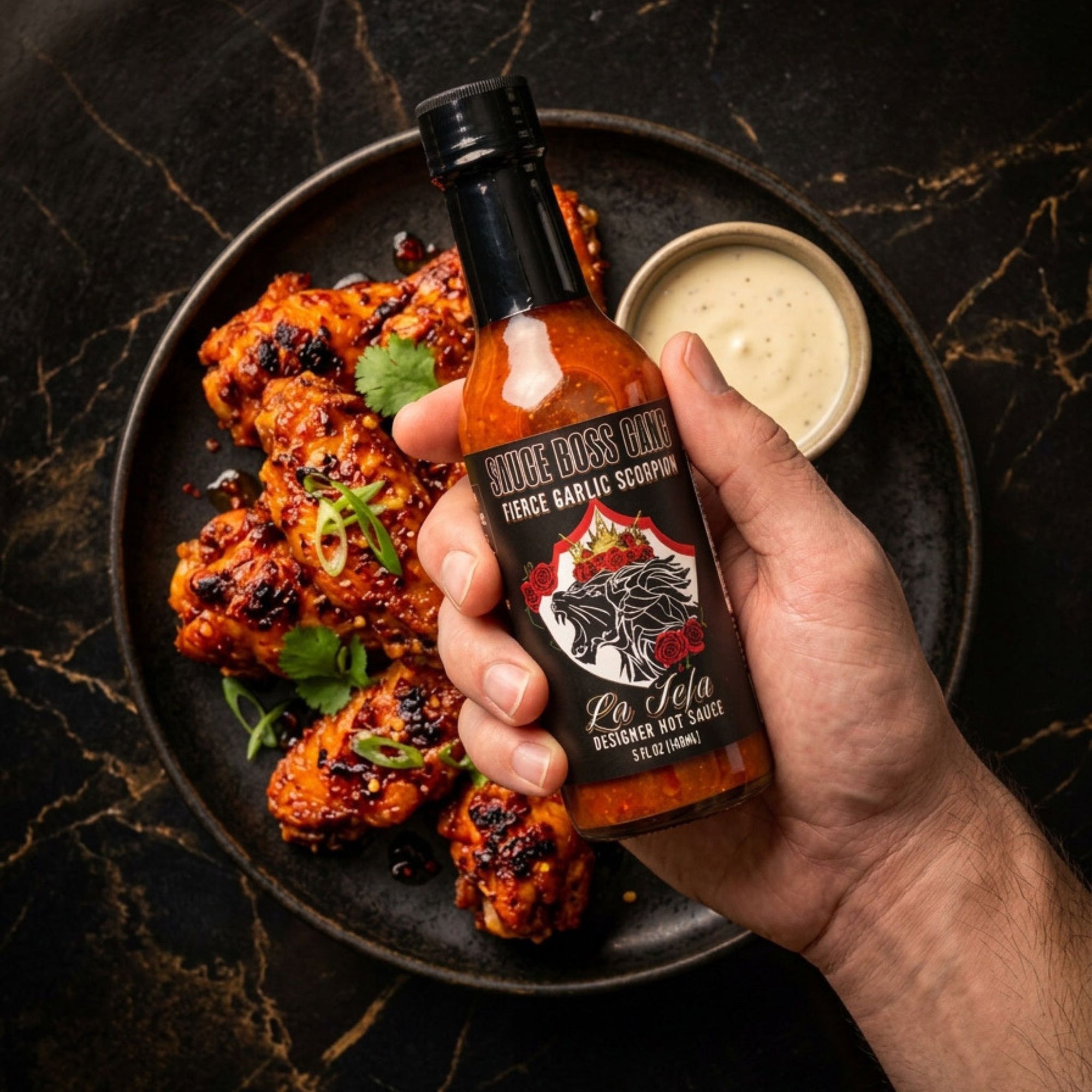 Hand holding a bottle of hot sauce over a plate of grilled chicken with a side of dipping sauce on a dark marble surface.