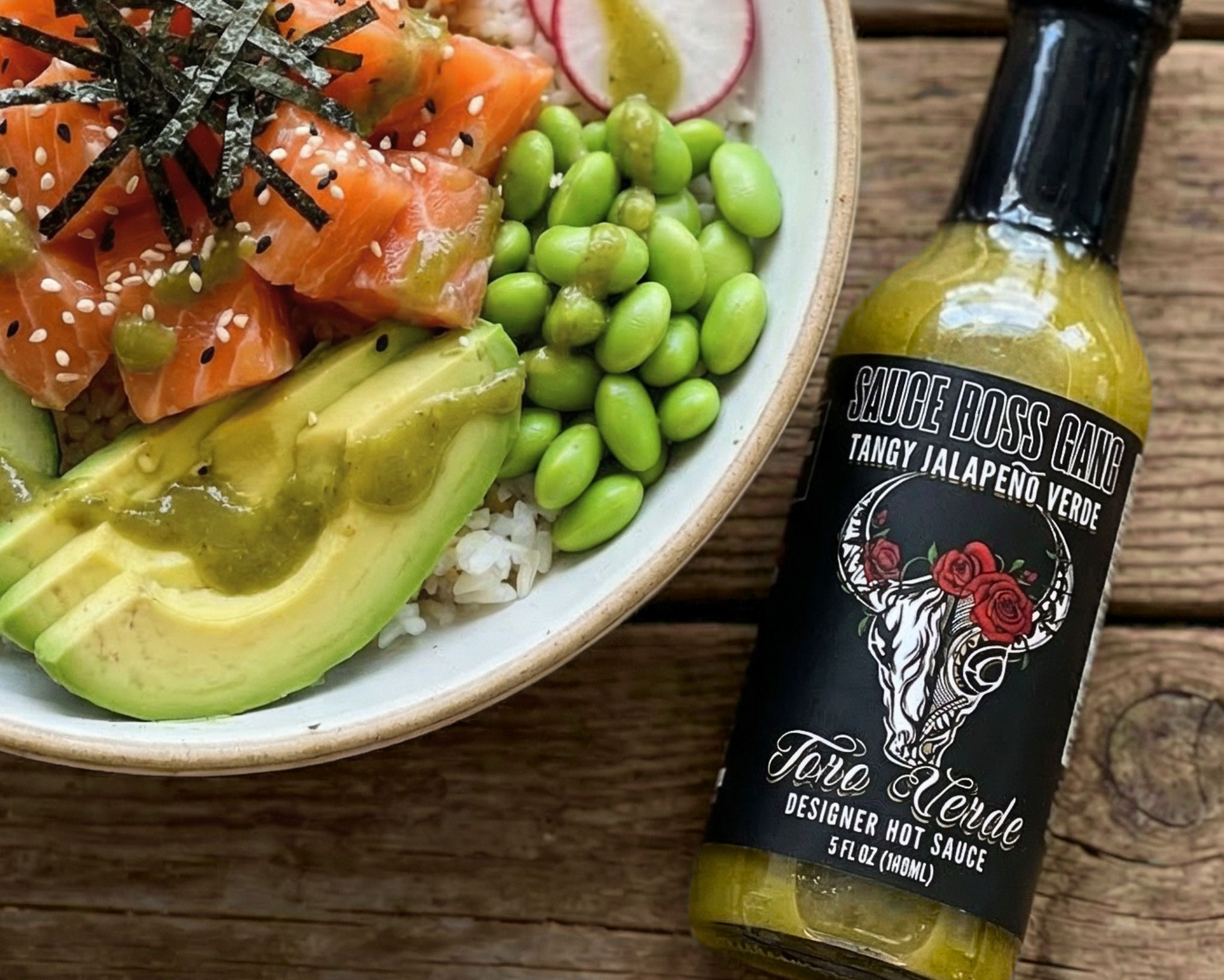 Bowl of sushi with salmon, avocado, and radish next to a bottle of 'Sauce Boss ' hot sauce on a wooden surface.