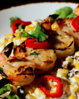 Plated dish of grilled shrimp and corn with red peppers and greens on a white plate.